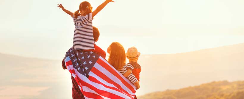 Família com pai, mãe e duas crianças em pé de costas vendo pôr do sol e segurando bandeira americana